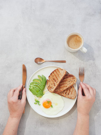 Hands holding cutlery, knife and fork, next to plate with toast, egg and avocado. Healthy breakfast with coffee.の写真素材