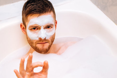 Young man with a facial mask lies in a foam in the bathroom. Concept of personal care for a man at...
