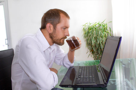 A man in white shirt drinking tea while sitting at the deskの写真素材