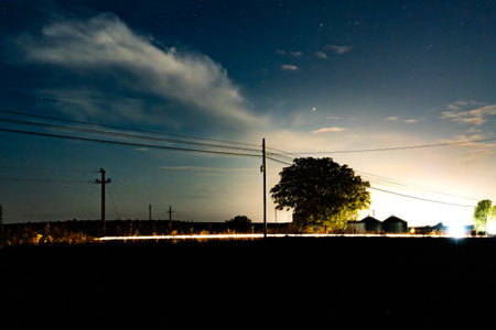 A rural area shows power lines in the night with clouds above. Distant lights can be seen near some houses. The field is dark, and stars are visible in the sky.の写真素材