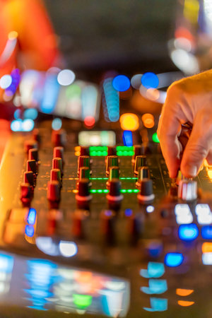 A DJ adjusts the sound levels on a mixer in a lively club. Bright lights shine in various colors...