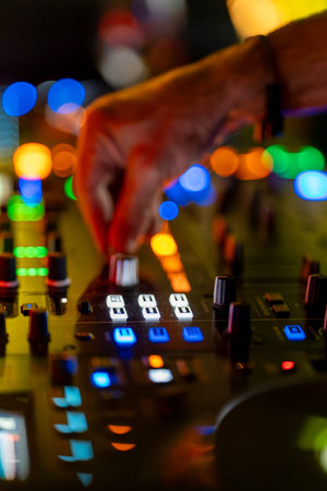A DJ is setting sound levels on a mixing console at a live music event. Bright lights of different...
