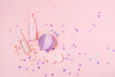 Flat lay composition with cosmetic bottle containers and bath bomb on pink color background with purple festive confetti.の写真素材
