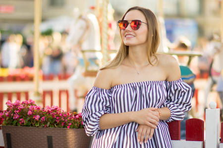 A girl in red sunglasses on vacation is smiling and enjoying a walk. Portrait of a beautiful young woman on a background of a bright merry-go-round. Lifestyle concept.の写真素材