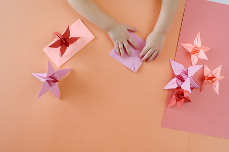 Children's hands do origami from colored paper on living coral background. Lesson of origami. Spring paper flower lie on a table. Top view, Flat lay styleの写真素材