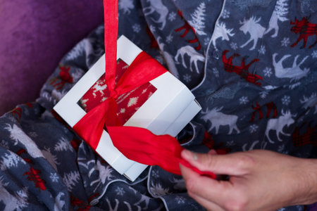 Man sitting in comfy pajamas opening Christmas present. Home holiday celebration. Classic white gift...