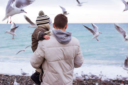 Father holding his son and feeding seagulls at the beach. Family time on the seaside. Man with boy feeds birdsの写真素材