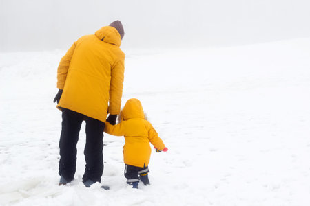 Father and son in warm yellow jacket back, outdoor family time, man holding hands with his child....