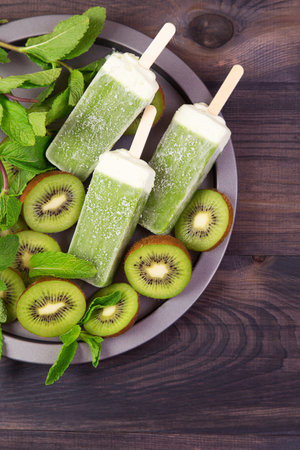 Homemade frosty kiwi ice sticks on a plate with mint and kiwi slices on a dark wooden background. Top view with copyspaceの写真素材