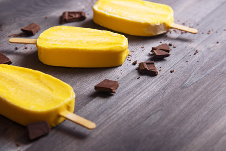 Yellow ice cream with chocolate bars and crumbs on dark wooden table. Space for text.