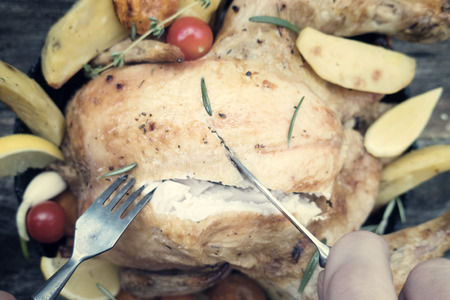 Appetizing baked chicken with vegetables on a wooden background and hands with a knife and fork that cut the chickenの写真素材
