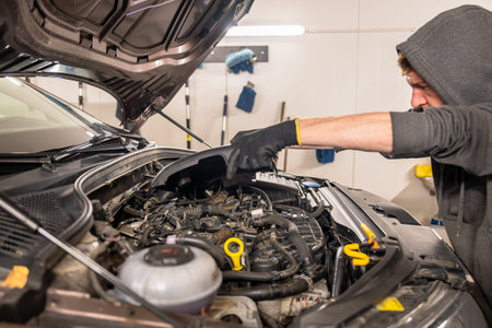 Mechanic wearing gloves and hoodie repairing a car engine in a well-lit garage, tools and equipment...