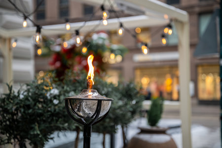 Close-up of an outdoor oil lamp with flame, string lights above, blurred plants, and a cozy urban background, evoking warmth, Christmas decorations.の写真素材