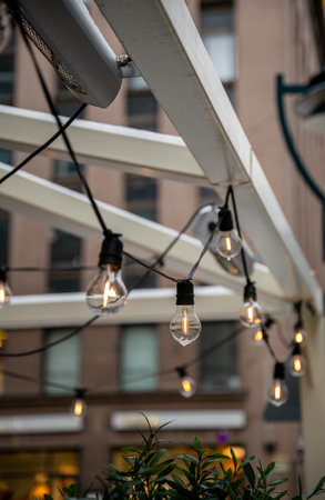 String lights hanging on a white pergola, with blurred urban building background and green foliage in foreground, cozy conceptの写真素材