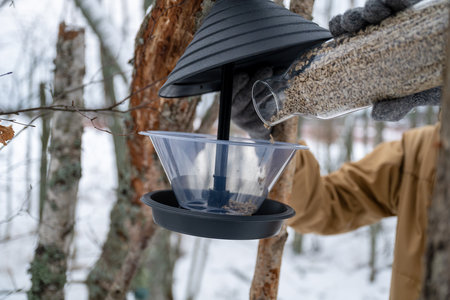 Person refilling a bird feeder attached to a tree in a snowy forest. concept of wildlife careの写真素材