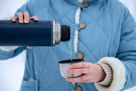 Close-up of hands pouring hot beverage from blue thermos into cup. Concept of warmth and outdoor...