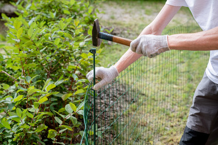 man in gloves hammering a fence stake into the ground in a lush garden setting, concept of outdoor workの写真素材