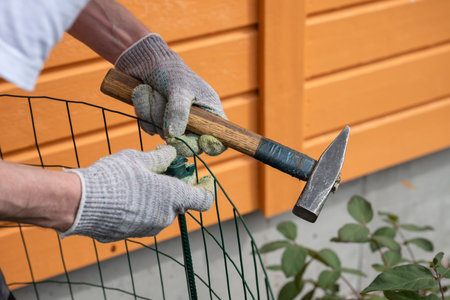 Person hammering green metal fence post with a steel hammer, wearing gloves. Concept of DIY or home repairの写真素材