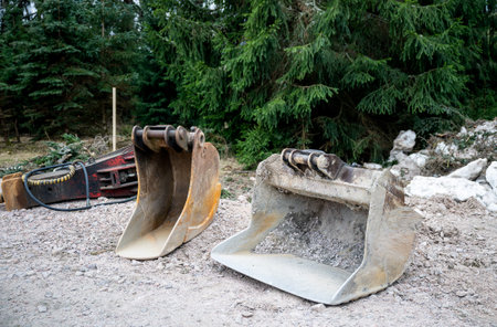 Two excavator buckets on a gravel surface with a hydraulic breaker, set against a forest background....