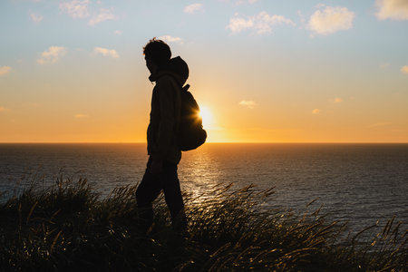 Silhouette of traveler walking on grassy coastline at sunset, Denmark