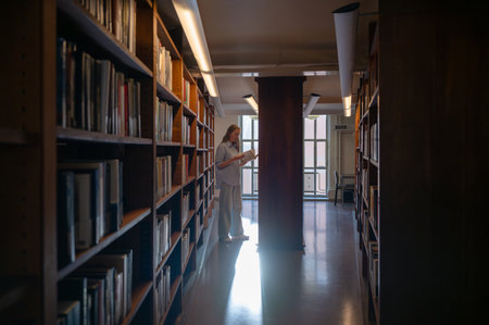 Woman reading a book in quiet library aisle with warm natural lightの写真素材
