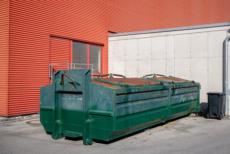 Industrial green metal dumpster near red and concrete building wall outdoors