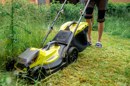 Man mowing overgrown lawn with yellow electric mower near wooden house stepsの写真素材