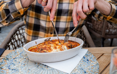Person cutting baked pasta Bolognese with fork and knife at outdoor restaurantの写真素材