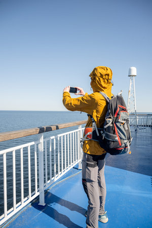 Back view of traveler in jacket taking smartphone photo on ferry deck at sea