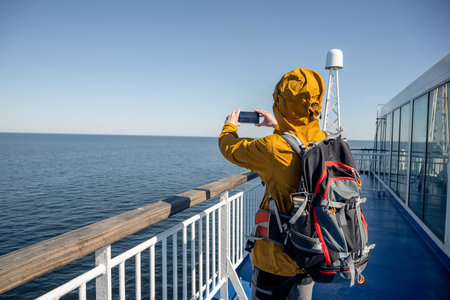 Back view of traveler in jacket taking smartphone photo on ferry deck at seaの写真素材