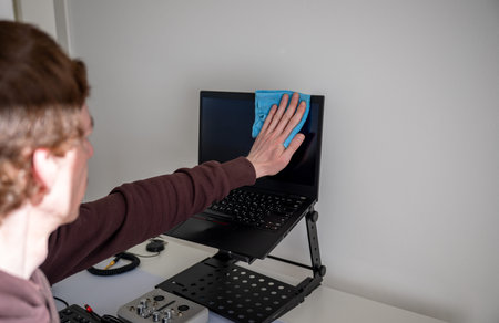 Person cleaning laptop screen with microfiber cloth on adjustable desk stand in home office