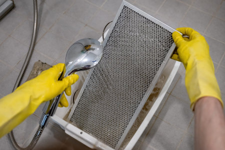 Person rinsing metal kitchen range hood filter with water in sink with yellow rubber gloves