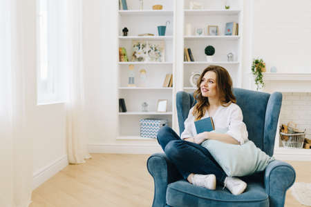 Young beautiful woman in an armchair with a book, relaxing in cozy living roomの写真素材