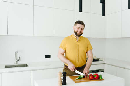 Handsome caucasian man in yellow cutting vegetables for meal or salad in modern white kitchen with a smileの写真素材