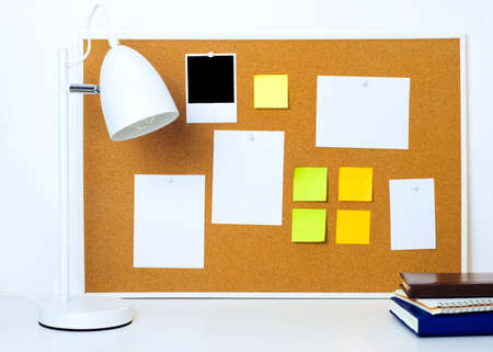 Framed cork notice board with various paper notes and stickers. Lamp and books on the foregroundの写真素材