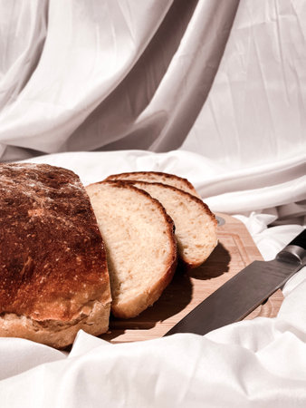 Freshly baked round bread cut into slices on a wooden board with a knife on a white fabric background. Close up.の写真素材