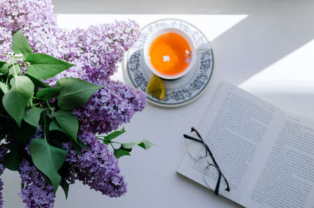 A top view of a lilac flower bouquet, cup of tea and a book on a window sill.の写真素材