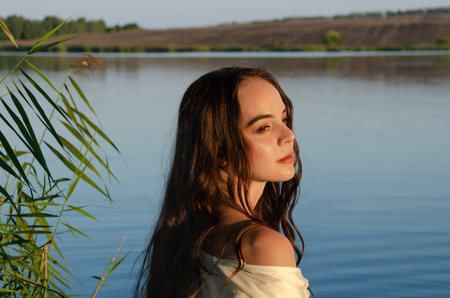 Young calm woman in white shirt stands on the lake reeds shore during sunset, with landscape behind...