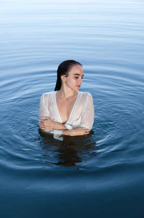 Young dreamy woman in white shirt standing in the lake, holding arms around herself, looking away.の写真素材