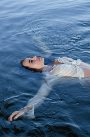 Beautiful young woman swimming in the lake with her arms spread in a white shirt.