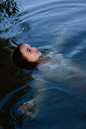 Beautiful young woman swimming in the lake with her arms spread in a white shirt.の写真素材