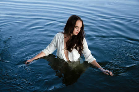 Young thoughtful brunette woman in white shirt is standing in the lake and moving hands through water.の写真素材