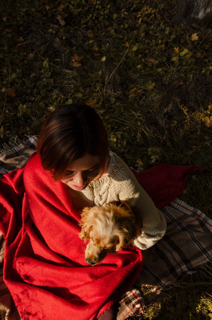 Top view of an unrecognisable woman sitting with her dog in autumn forest, cuddling together in red blanket.の写真素材
