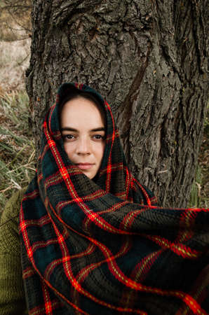 Closeup portrait of a brunette woman standing next to the tree, wrapped in a tartan scarf, looking at the camera.の写真素材