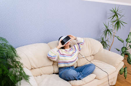 Full body shot of a teen girl in VR glasses sitting on a couch in her room. Entertainment, cyberspace concept.の写真素材