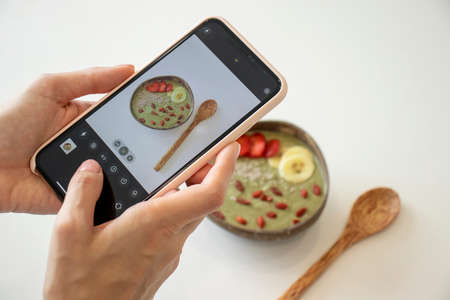 A young woman photographs a bowl of green smoothie with fruit and goji berries. Top viewの写真素材