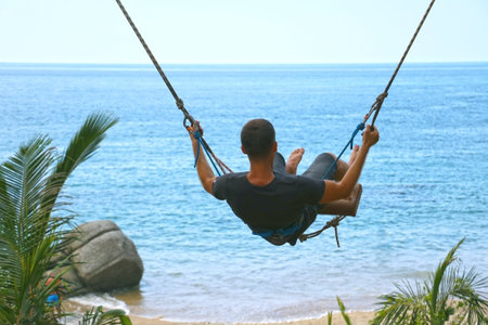 Young man riding a swing high in the sky against a background of ocean and sea. Guy swinging view...
