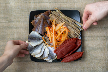 Dried Seafood Mix with Squid, Tuna, codfishon, yellow minke on Plate Top View. Cured Dehydrated Salty Fish Fillet as Elite Beer Snack. Mans hand and a womans hand take the fish.の写真素材