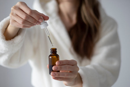 Young woman in a bathrobe is holding a bottle with a dropper of oil or serum for face and body care.の写真素材