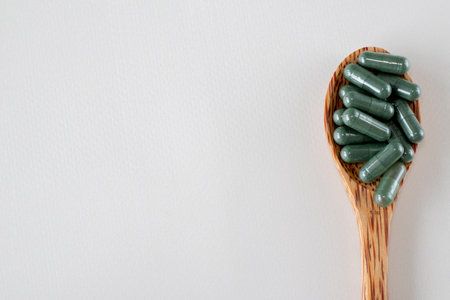 Dark green capsules with chlorophyll or spirulina. Useful supplements or medicines in a wooden spoon on a beige background. Copy space for textの写真素材
